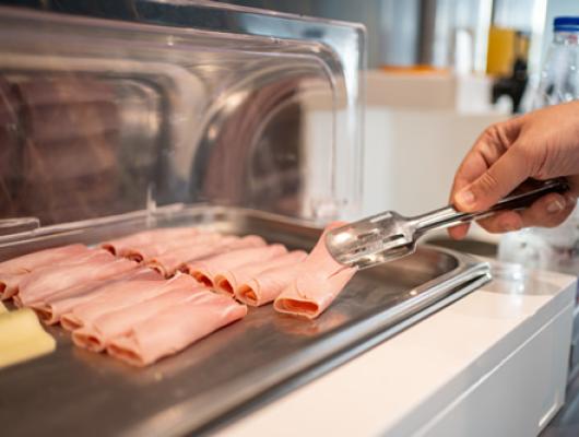 Person taking slices of ham with tongs in a cafeteria.