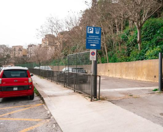 Parcheggio riservato con cancello, auto rosse e vegetazione sullo sfondo.