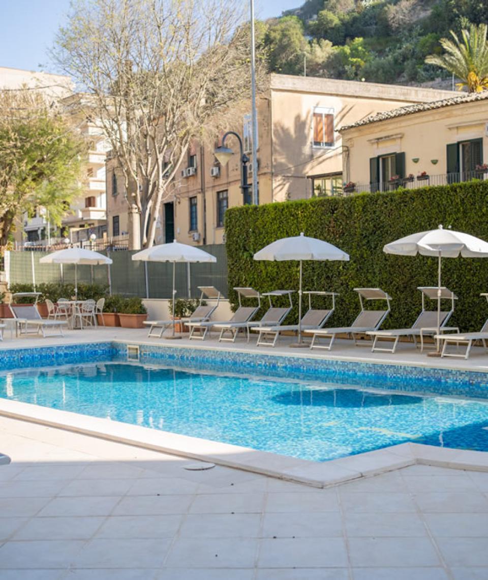 Outdoor pool with sunbeds and umbrellas, surrounded by buildings and greenery.