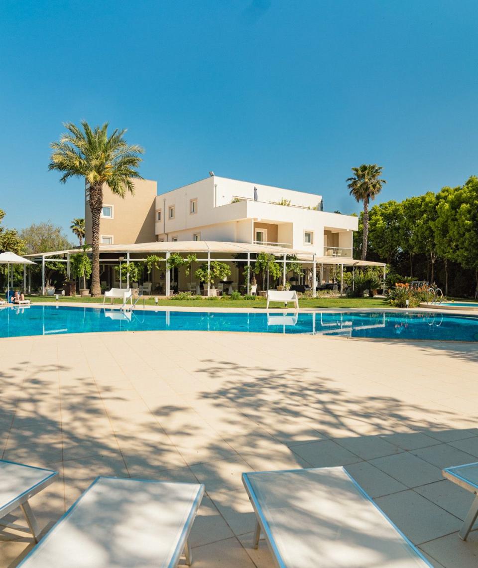 Outdoor pool with sun loungers, umbrellas, and palm trees in a sunny setting.